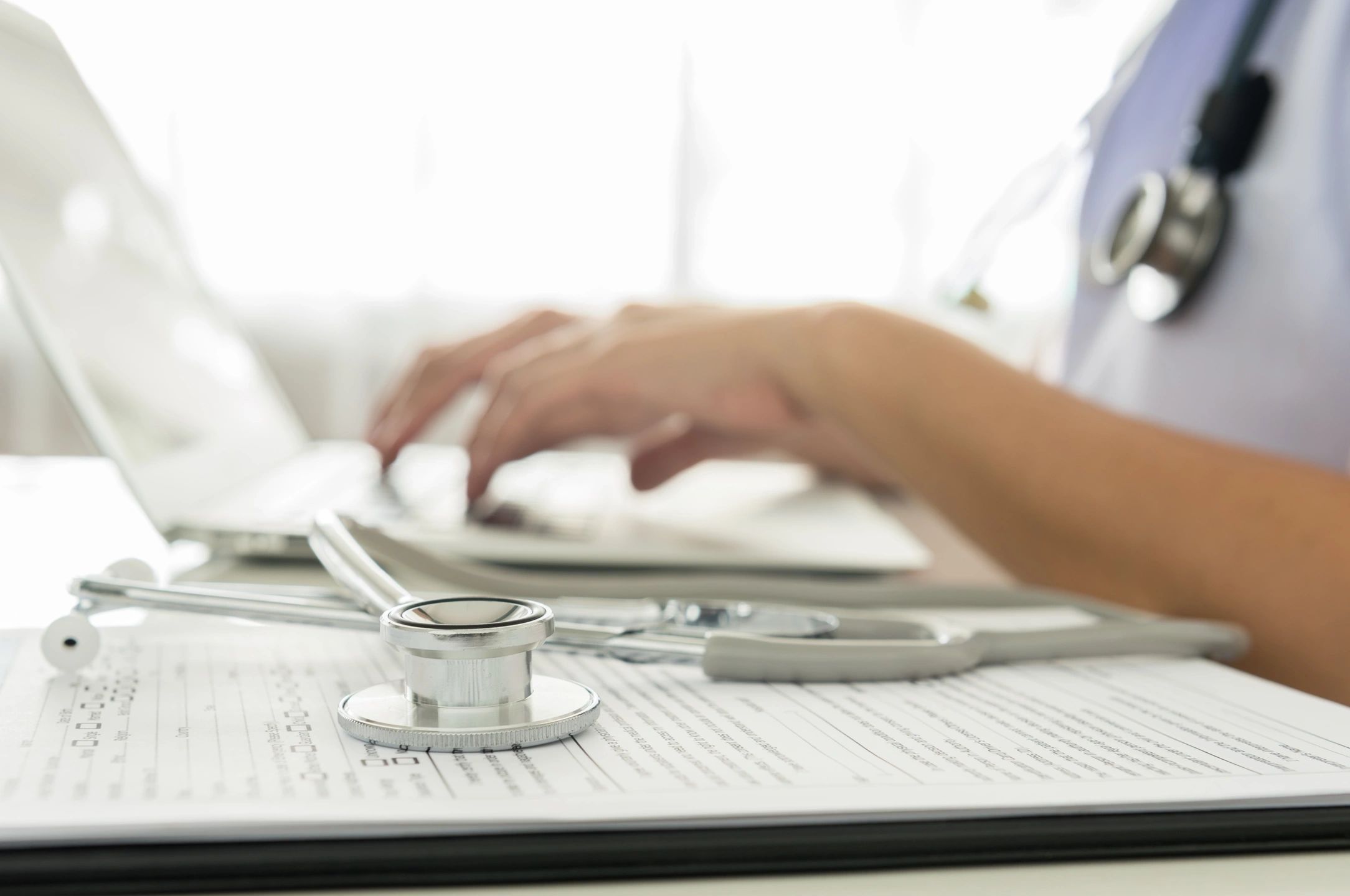 nurse typing on computer with stethoscope laying next to it on a notebook